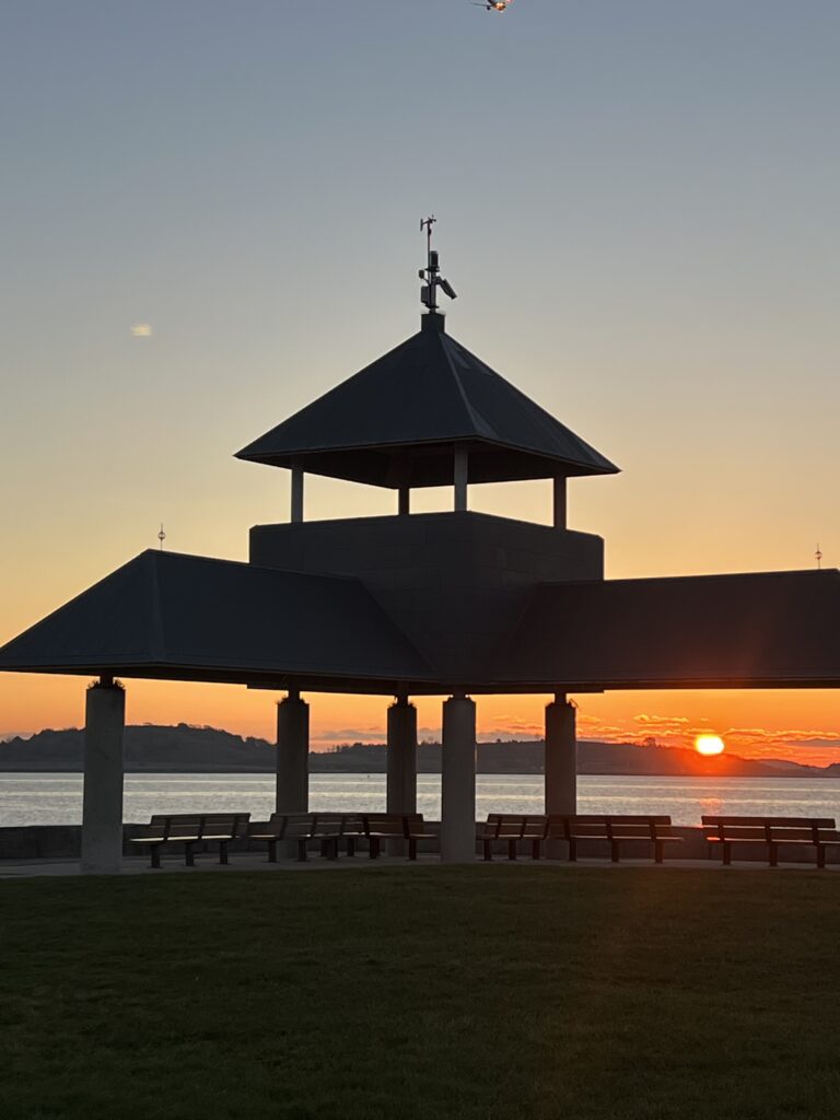 A low-angle shot captures the dark silhouette of the Pleasure Bay Pavilion at Castle Island in South Boston, set against a vibrant sunset over Boston Harbor.