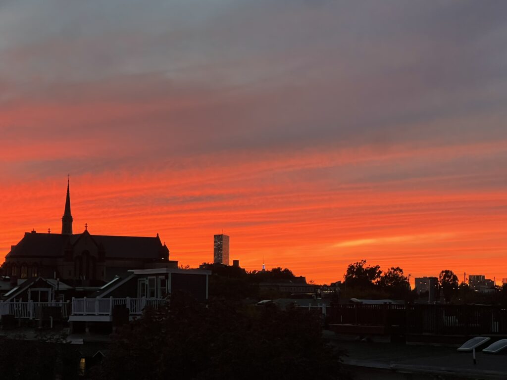 A wide, eye-level shot captures a silhouette of the Boston skyline against a dramatic sunset filled with fiery red and orange clouds.