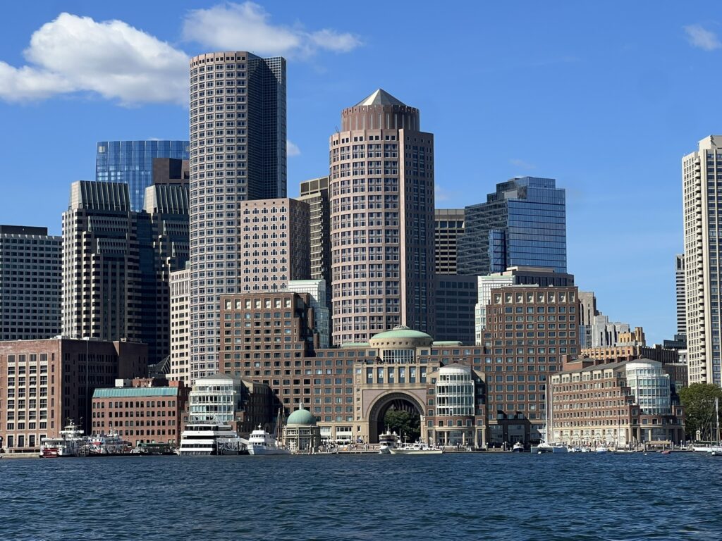 A wide, eye-level shot captures a dense cluster of skyscrapers in downtown Boston, Massachusetts, as seen from across the blue waters of Boston Harbor under a clear blue sky with a few faint white clouds.
