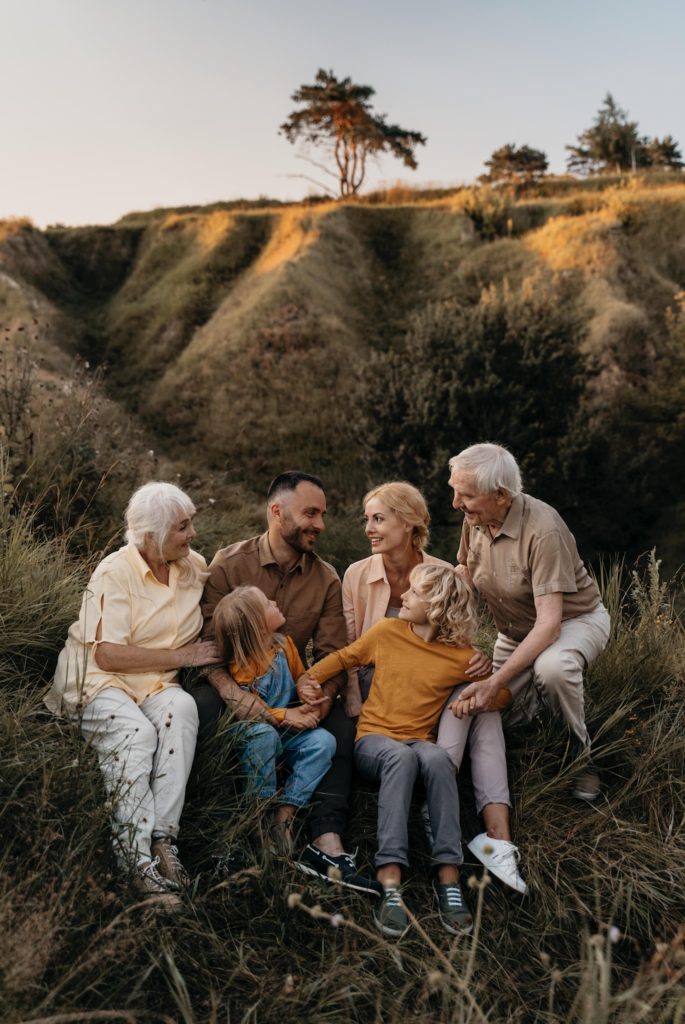 A medium, eye-level shot captures a multigenerational family of six sitting together in tall, wild grass on a rustic, sloping hillside during golden hour.